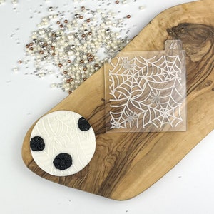 May include: A white fondant cake round with a black spiderweb design and three black spiders. A clear plastic stencil with a white spiderweb design and three spiders is also shown.
