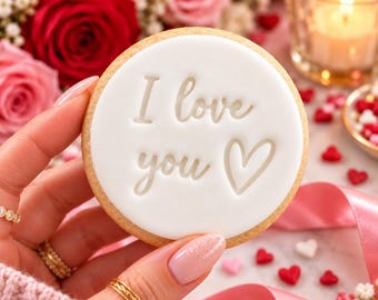 Puede incluir: Una galleta redonda con glaseado blanco liso que presenta el texto en relieve I love you y un símbolo de corazón. La galleta tiene un borde dorado y se sostiene sobre un fondo de rosas rojas y rosas, una vela encendida y chispas de corazón.