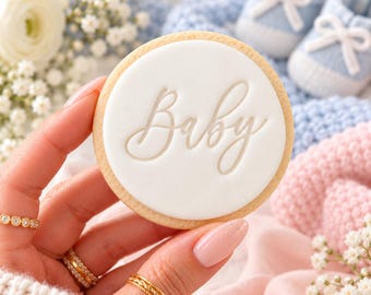 May include: A round sugar cookie topped with smooth white fondant featuring the embossed word Baby in a cursive script font. The cookie is held against a backdrop of soft blue and pink knitted fabric, baby booties, and white flowers.