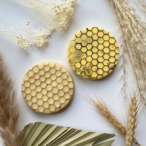 Peut inclure: Deux biscuits ronds décorés d'un motif en nid d'abeille. Un biscuit est jaune avec des lignes noires et l'autre est blanc avec des lignes dorées.