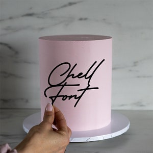 May include: A pink cake with black cursive writing that says "Chell Font" on the side.