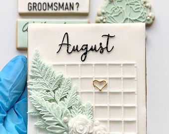 May include: White square cookie decorated with a green fern, white flowers, and a gold heart on a white grid. The cookie is labeled "August" in black cursive.