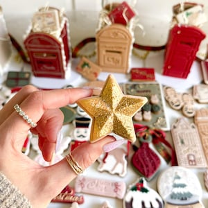 May include: A gold star-shaped cookie with glitter detailing is held in front of a collection of Christmas-themed cookies. Other cookies include red mailboxes, ornaments, and snow globes. The cookies are decorated with icing and various festive designs.