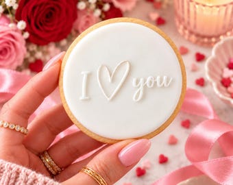 Puede incluir: Una galleta redonda con glaseado blanco liso que presenta el texto en relieve blanco I heart you. La galleta tiene un borde dorado y se sostiene sobre un fondo de rosas rojas y rosas, una vela encendida y pequeños confites de corazón.