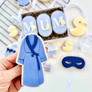 May include: A light blue robe-shaped cookie held in a hand, surrounded by other cookies. The cookies include a sleep mask, rubber duck, and cookies spelling "MUM". The cookies are decorated with blue and yellow icing.