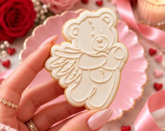 May include: A light-colored sugar cookie shaped like a teddy bear with angel wings, featuring embossed details of a bear holding a heart. The cookie has a smooth, matte finish and is held up against a soft, light-toned background.