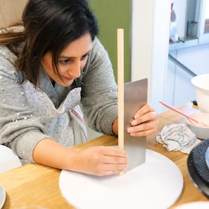 May include: A stainless steel cake scraper held against a vertical wooden dowel on a white circular cake board. The scraper is rectangular with a flat edge, and the dowel is cylindrical and light-coloured, positioned upright on the board.