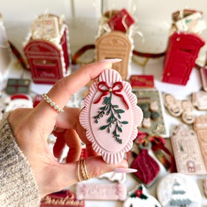May include: A pink, oval-shaped cookie with a scalloped edge, decorated with a red bow and green mistletoe. Other Christmas-themed cookies and decorations are in the background, including red mailboxes and gift boxes.