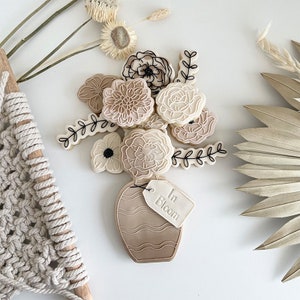 May include: A set of decorated sugar cookies in the shape of flowers and a vase. The cookies are in shades of brown and white with black outlines. The vase has a tag that says "In Bloom".