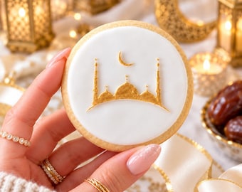 May include: A round sugar cookie with a smooth white icing surface featuring a gold-colored silhouette of a mosque with minarets and a crescent moon centered above the domes, held up against a blurred background of gold lanterns.