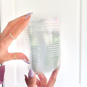 May include: A stack of clear, rectangular plastic containers, held by a person. The containers are transparent, allowing a glimpse of the background. The person's hands are visible, adorned with rings and light purple nail polish.