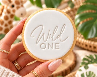 Może przedstawiać: Okrągłe ciastko z gładką białą warstwą fondantu z wytłoczonym napisem Wild ONE wykonanym połączeniem pisma odręcznego i czcionki bezszeryfowej. Ciastko ma złocistobrązowy brzeg i jest trzymane w dłoni.