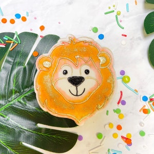 May include: A decorated sugar cookie shaped like a lion's face. The cookie is orange with gold glitter and has a white face with black eyes and a black nose. The cookie is surrounded by colourful sprinkles.