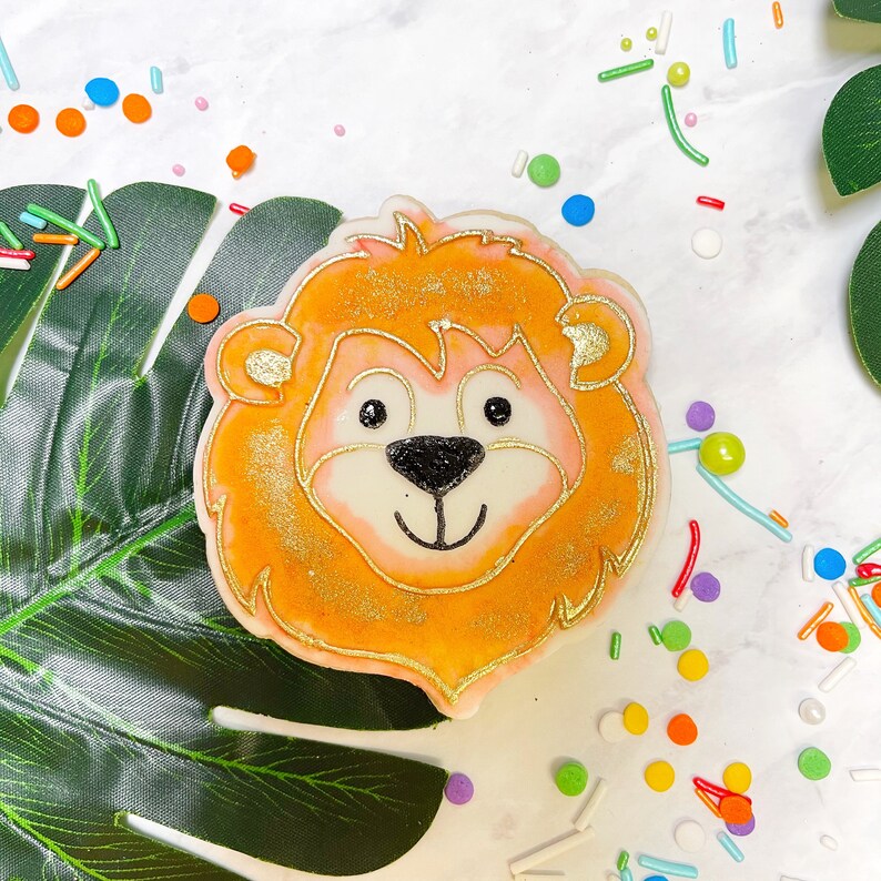 May include: A decorated sugar cookie shaped like a lion's face. The cookie is orange with gold glitter and has a white face with black eyes and a black nose. The cookie is surrounded by colourful sprinkles.