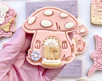 Op de afbeelding: Een roze en witte suikerkoekje versierd om eruit te zien als een paddenstoelenhuis. De koek heeft een witte deur met gouden accenten en een klein paars bloemetje.