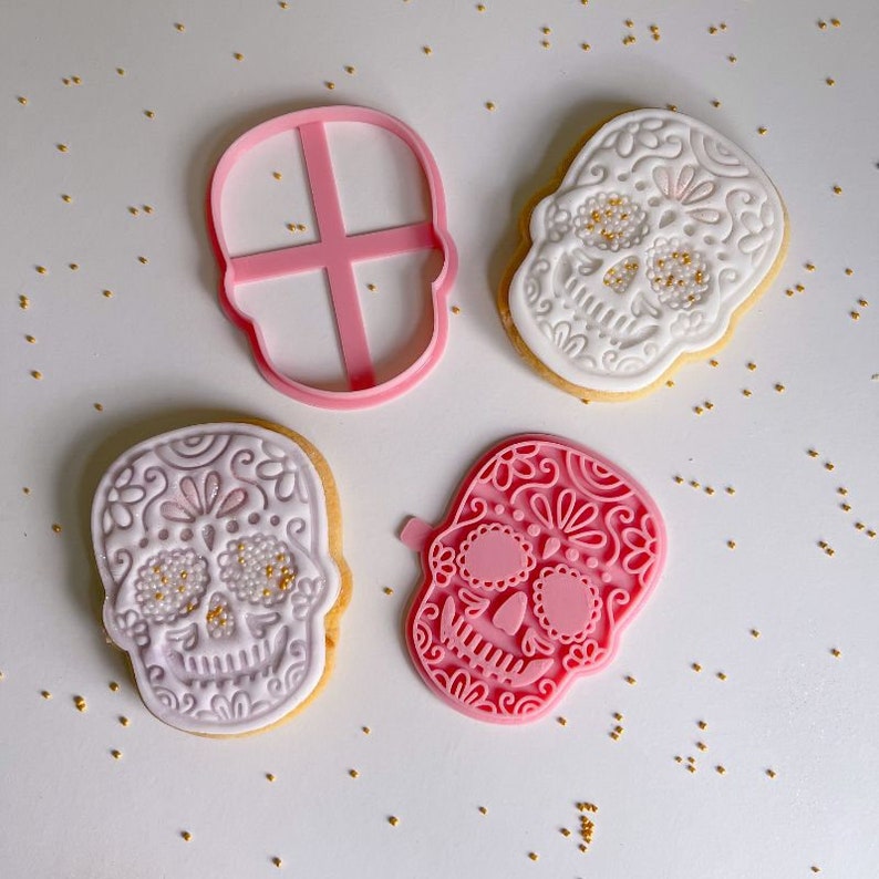 May include: Two sugar skull cookies decorated with white and light purple icing and gold sprinkles, alongside a pink plastic skull-shaped cookie cutter and a matching pink plastic skull-shaped cookie stamp with intricate floral patterns.