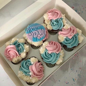 K&ouml;nnte beinhalten: Eine Schachtel mit sechs Cupcakes, die mit blauem und rosa Frosting und wei&szlig;en Streuseln verziert sind. Die Cupcakes haben ein Gender-Reveal-Thema mit einem blauen und wei&szlig;en Cupcake mit einem blauen Zuckerguss-Topper, auf dem "Er oder Sie?" steht.