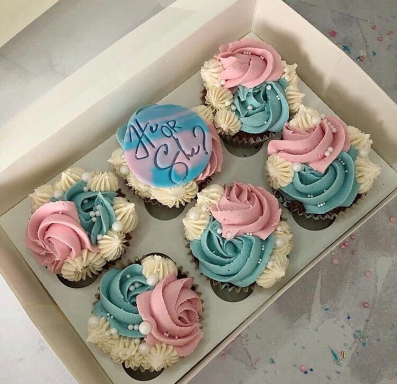 K&ouml;nnte beinhalten: Eine Schachtel mit sechs Cupcakes, die mit blauem und rosa Frosting und wei&szlig;en Streuseln verziert sind. Die Cupcakes haben ein Gender-Reveal-Thema mit einem blauen und wei&szlig;en Cupcake mit einem blauen Zuckerguss-Topper, auf dem "Er oder Sie?" steht.