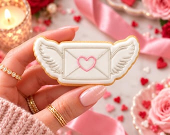 May include: A rectangular sugar cookie shaped like a winged envelope, featuring white icing, silver piped outlines, and a central pink heart. The cookie is held horizontally, showcasing detailed feathered wings on both sides of the envelope.