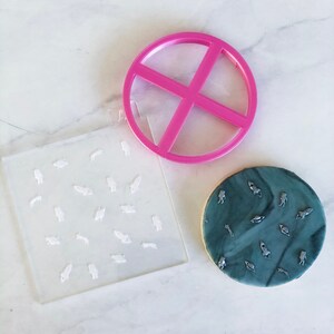 Pode incluir: Um conjunto de cortadores de biscoitos para decorar biscoitos. O conjunto inclui um cortador redondo com um design de cruz, um estêncil de plástico transparente com um padrão de pequenos foguetes e astronautas, e um biscoito decorado com glacê marmorizado azul e cinza e decorações de açúcar prateadas.