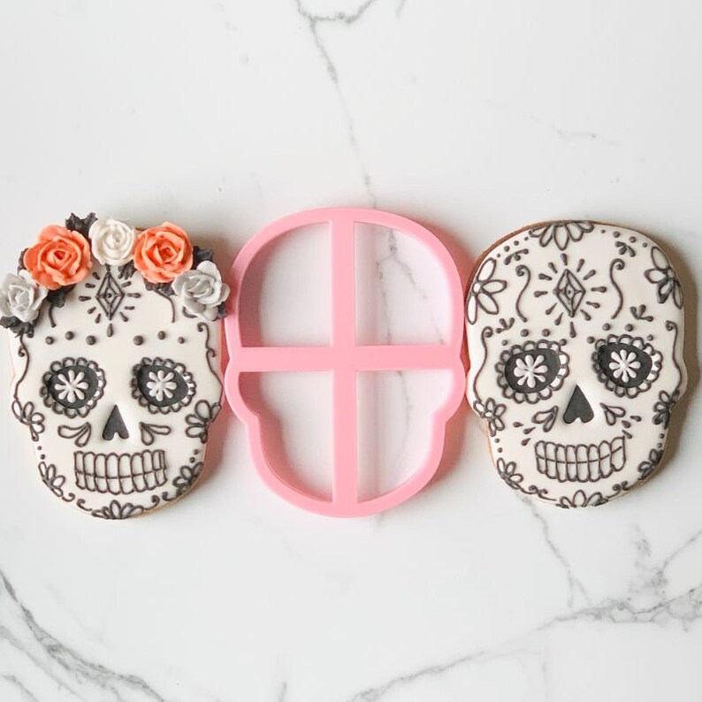 May include: Two sugar skull cookies decorated with white icing, black intricate patterns, and orange and white floral accents, positioned on either side of a pink plastic skull-shaped cookie cutter with a central cross-shaped support bar.