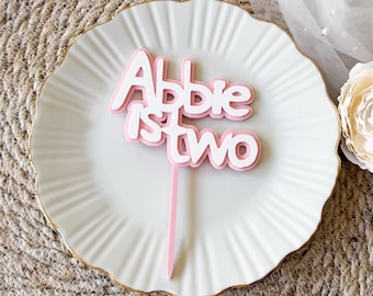 Puede incluir: Un adorno para pastel rosa y blanco que dice "Abbie is two". El adorno es en capas, con una base rosa y letras blancas. Se coloca sobre un plato blanco con borde dorado, con un fondo neutro.