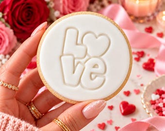 Puede incluir: Una galleta redonda con glaseado blanco liso que presenta la palabra LOVE en relieve, donde la letra O es reemplazada por una forma de corazón. La galleta tiene un borde dorado y se sostiene sobre un fondo de rosas rojas y cinta rosa.