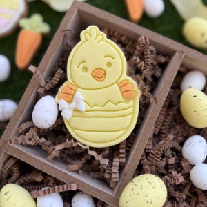 Puede incluir: Una galleta de pollito amarillo decorada con detalles naranjas y blancos, sentada en una caja de madera llena de papel marrón con otras galletas de temática de Pascua.
