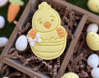 Op de afbeelding: Een gele kuikenkoekje versierd met oranje en witte details, in een bruine, met papier gevulde houten doos met andere paaskoekjes.