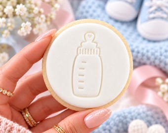 May include: A round sugar biscuit topped with a smooth white fondant layer featuring an embossed outline of a baby bottle with a small heart detail on the nipple. The biscuit has a light golden-brown edge and a clean, flat surface design.