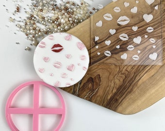 May include: A white fondant cake decorated with pink hearts and red lips. A clear plastic stencil with hearts and lips is shown next to the cake. A pink plastic circle cutter with four sections is also shown.