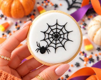 May include: A round sugar cookie with a smooth white icing base, featuring a black piped spider web design and a small black spider. The cookie has a light golden-brown edge and is held against a background of pumpkins and candy.