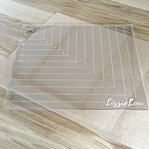 Peut inclure: Un tapis de coupe en acrylique transparent avec des mesures en pouces de 2 à 14. Le tapis a une grille et est étiqueté "Lissle Lou".