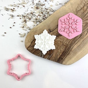 Puede incluir: Tres cortadores de galletas con forma de copo de nieve, uno rosa, uno blanco y uno rosa con un diseño de copo de nieve detallado. Los cortadores están sobre una superficie de madera.