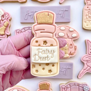 May include: A pink and gold cookie shaped like a bottle with a cork top. The cookie is decorated with gold stars and the words "Fairy Dust" on a white label.