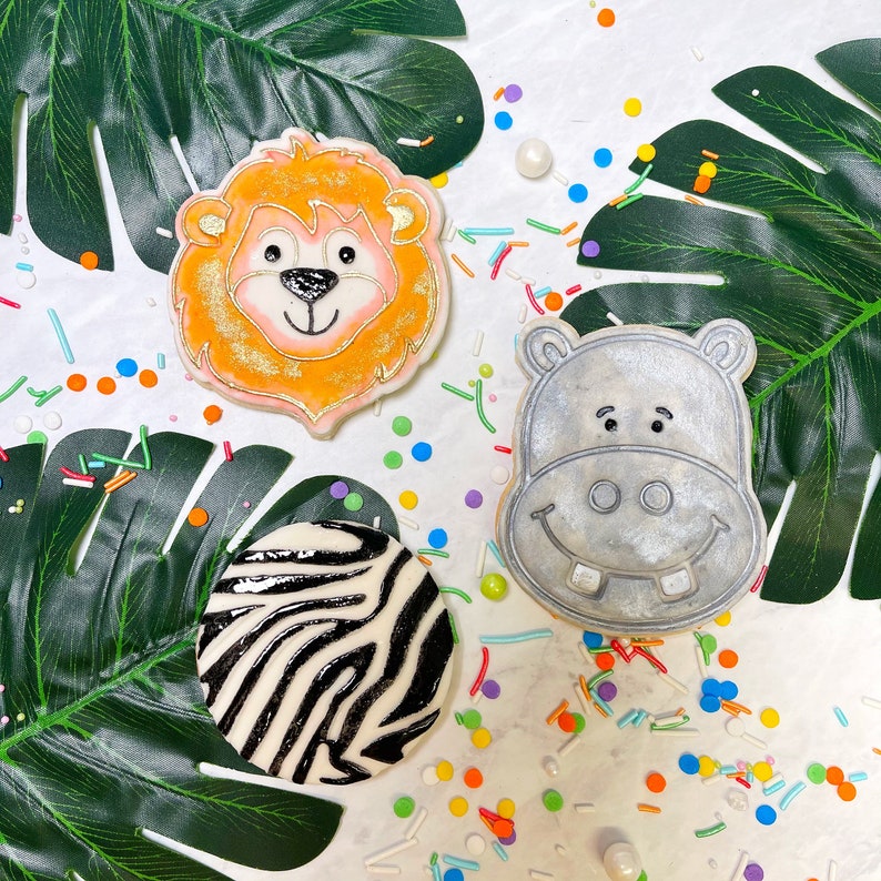 May include: Three decorated sugar cookies on a white background with colourful sprinkles. The cookies are shaped like a lion, a hippopotamus, and a zebra. The lion cookie is orange with a brown mane and a black nose. The hippopotamus cookie is grey with a black nose and mouth. The zebra cookie is black and white.
