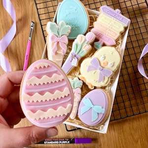 Puede incluir: Una caja de galletas con temática de Pascua, que incluye formas de huevo, zanahoria y pollito, decoradas con colores pastel y lazos. Las galletas tienen las palabras "Happy Easter" y "Carrot Patch". También se ve un bolígrafo morado y un pincel.