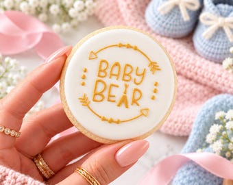 Könnte beinhalten: Ein runder Keks mit weißem Zuckerguss und goldener Aufschrift BABY BEAR, verziert mit goldenen Pfeilen und Punkten. Der Keks wird vor hellblauen gehäkelten Babyschuhen und einem rosa Stoff präsentiert.