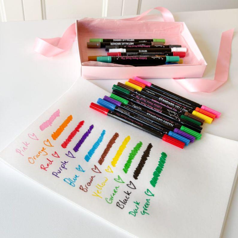 Puede incluir: Un juego de rotuladores comestibles de doble punta en una caja rosa claro con cinta, acompa&ntilde;ado de una hoja blanca que muestra muestras de color y etiquetas escritas a mano como Pink, Orange, Red, Purple, Blue, Brown, Yellow, Green, Black y Dark Green.