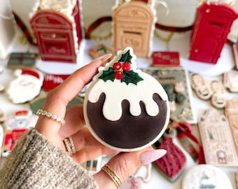 Pode incluir: Um biscoito de pudim de Natal com glacê branco e bagas de azevinho é segurado em frente a outros biscoitos com tema natalino. Outros biscoitos incluem uma caixa de correio vermelha, um Papai Noel e uma bola de neve. Os biscoitos são decorados com glacê vermelho, branco, verde e marrom.