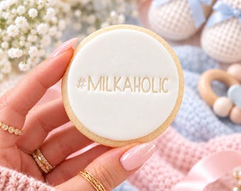 May include: A round sugar cookie with a smooth white fondant topping featuring the embossed text #MILKAHOLIC. The cookie is held against a soft background of baby blue and pink knitted textures, white baby booties, and flowers.