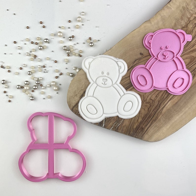 May include: Two pink plastic teddy bear cookie cutters and one white embossed teddy bear cookie shape. The cutters feature a seated bear design with rounded ears, arms, and legs. Small metallic and white pearl sprinkles are scattered nearby.