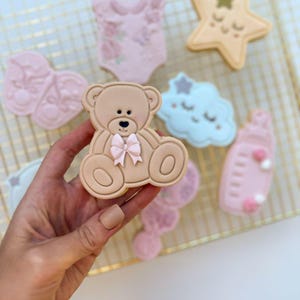 May include: A beige teddy bear-shaped cookie with a small pink bow at the neck, held against a background of other baby-themed cookies including a star, a cloud, a baby bottle, and a onesie, all decorated with pastel icing on a wire rack.