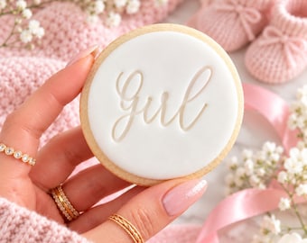May include: A round sugar cookie with smooth white icing featuring the word girl embossed in a cursive script font. The cookie is held against a soft pink knitted fabric background with small white flowers and tiny pink baby booties.