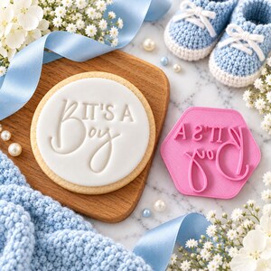 May include: A round cookie with white icing embossed with the text It's a Boy, accompanied by a pink hexagonal plastic stamp featuring the same text, light blue crocheted baby booties, white flowers, and a light blue satin ribbon.