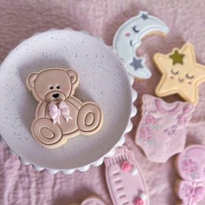 May include: A light brown teddy bear cookie with a pink bow sits on a white speckled plate. Nearby are cookies shaped like a crescent moon, a star, a baby onesie with floral patterns, a baby bottle, and a rattle, all decorated in soft pastel tones.