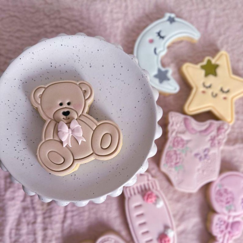 May include: A light brown teddy bear cookie with a pink bow sits on a white speckled plate. Nearby are cookies shaped like a crescent moon, a star, a baby onesie with floral patterns, a baby bottle, and a rattle, all decorated in soft pastel tones.
