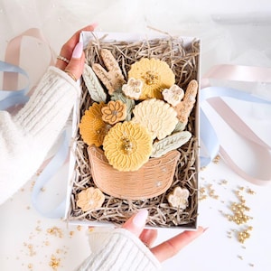 Könnte beinhalten: Eine weiße Geschenkbox gefüllt mit braunem zerkleinertem Papier und verzierten Zuckerkeksen in Form von Blumen in einem Korb. Die Kekse sind gelb, weiß und grün.