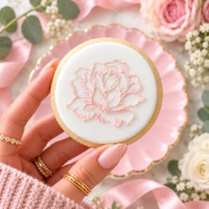 Può includere: Un biscotto rotondo con una base di glassa bianca liscia caratterizzato da un delicato disegno a contorno di una rosa rosa chiaro disegnato a mano. Il biscotto ha un bordo dorato e viene tenuto su uno sfondo chiaro.