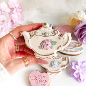Può includere: Un biscotto a forma di teiera color crema decorato con una rosa rosa e foglie verdi. Il biscotto ha accenti dorati ed è circondato da altri biscotti e fiori. Il biscotto è tenuto da una persona.